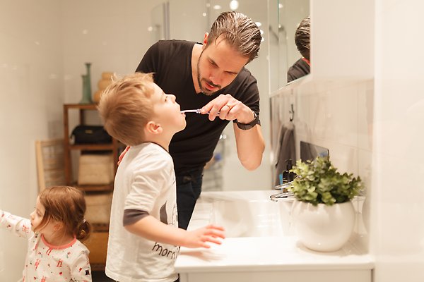 En pappa borstar tänderna på sin son inne på toaletten. Ett mindre barn syns i bakgrunden.