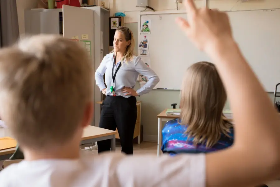 En lärare står i ett klassrum omgiven av elever som räcker upp handen. 
